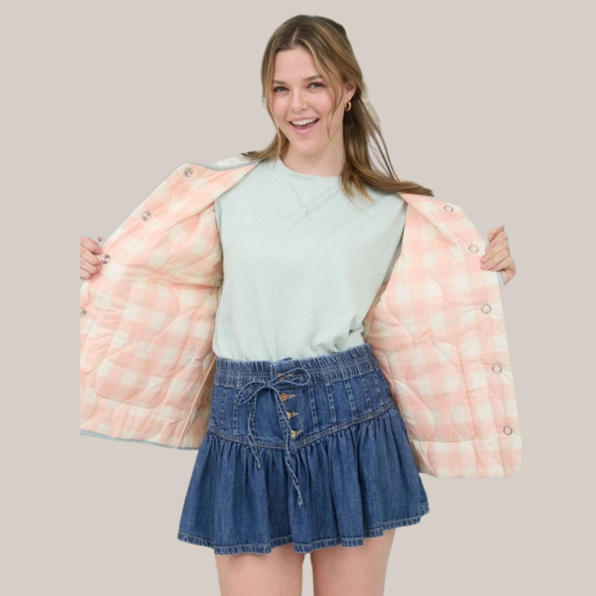 Woman wearing a light pink checkered jacket over a light blue top and denim skirt on a beige background