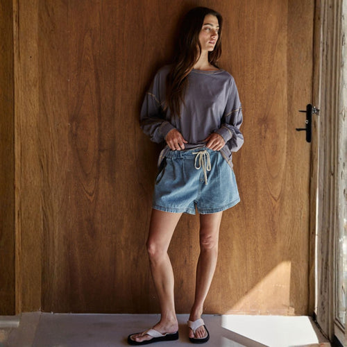 Woman wearing a gray sweatshirt and denim shorts standing against a wooden wall.